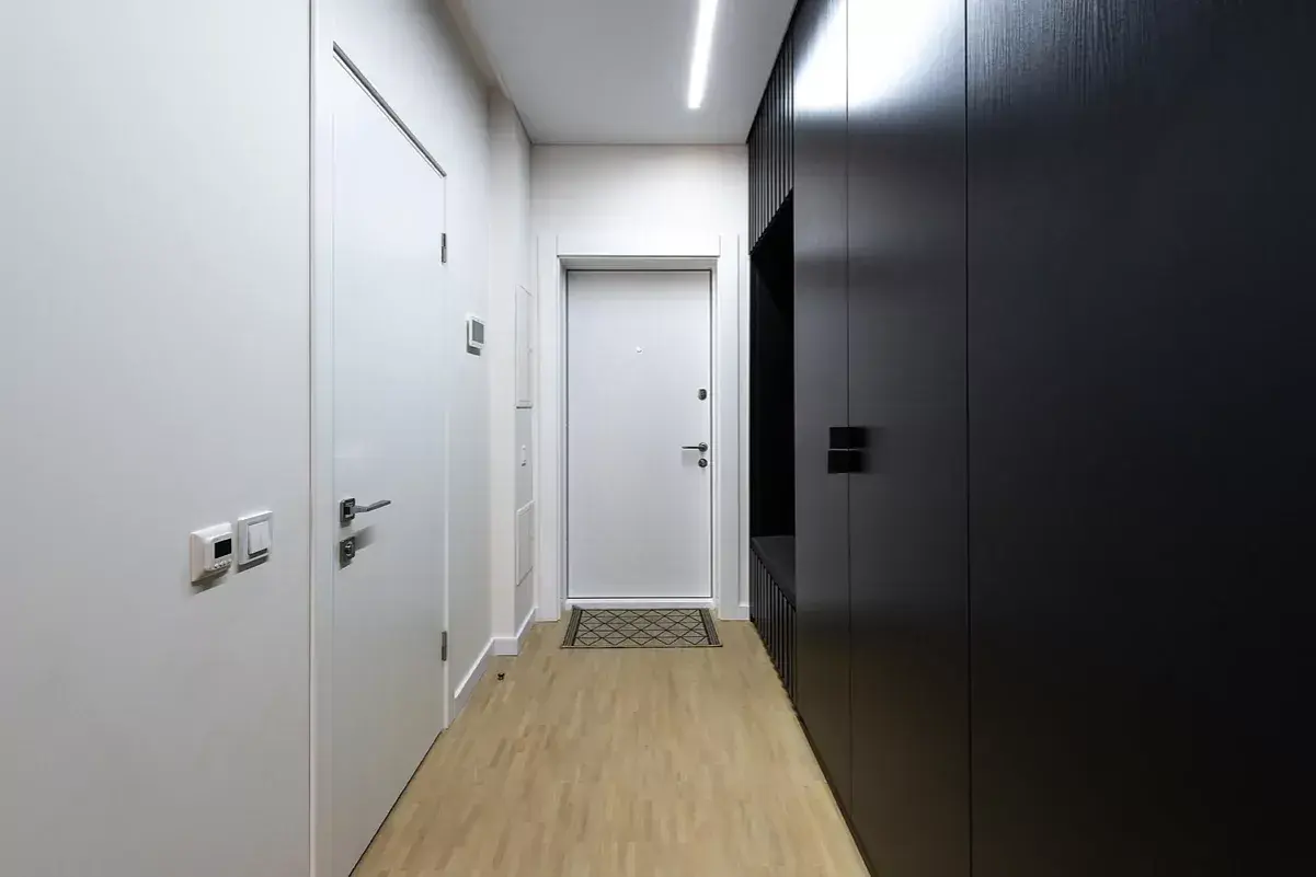 Pasillos estrechos y modernos con puertas blancas y un armario negro. El suelo de madera clara y la iluminación lineal crean un ambiente minimalista.
