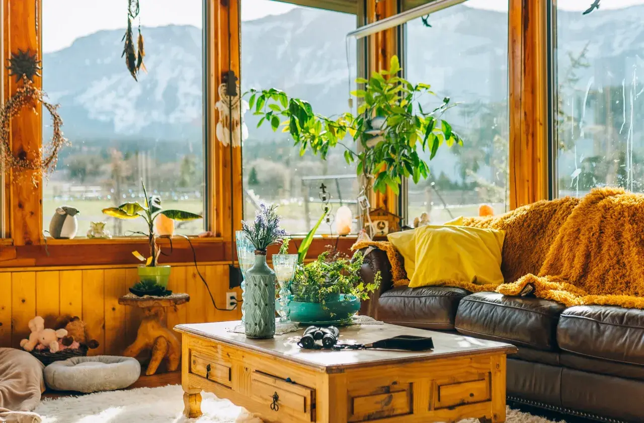 Un salón acogedor con sofá de cuero, manta naranja, cojín amarillo y mesa de centro de madera. Grandes ventanales con vistas a la montaña.