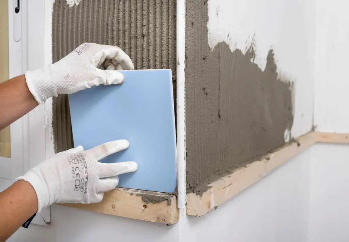 Manos enguantadas colocando un azulejo azul sobre cemento fresco. Guía visual de como poner azulejos.
