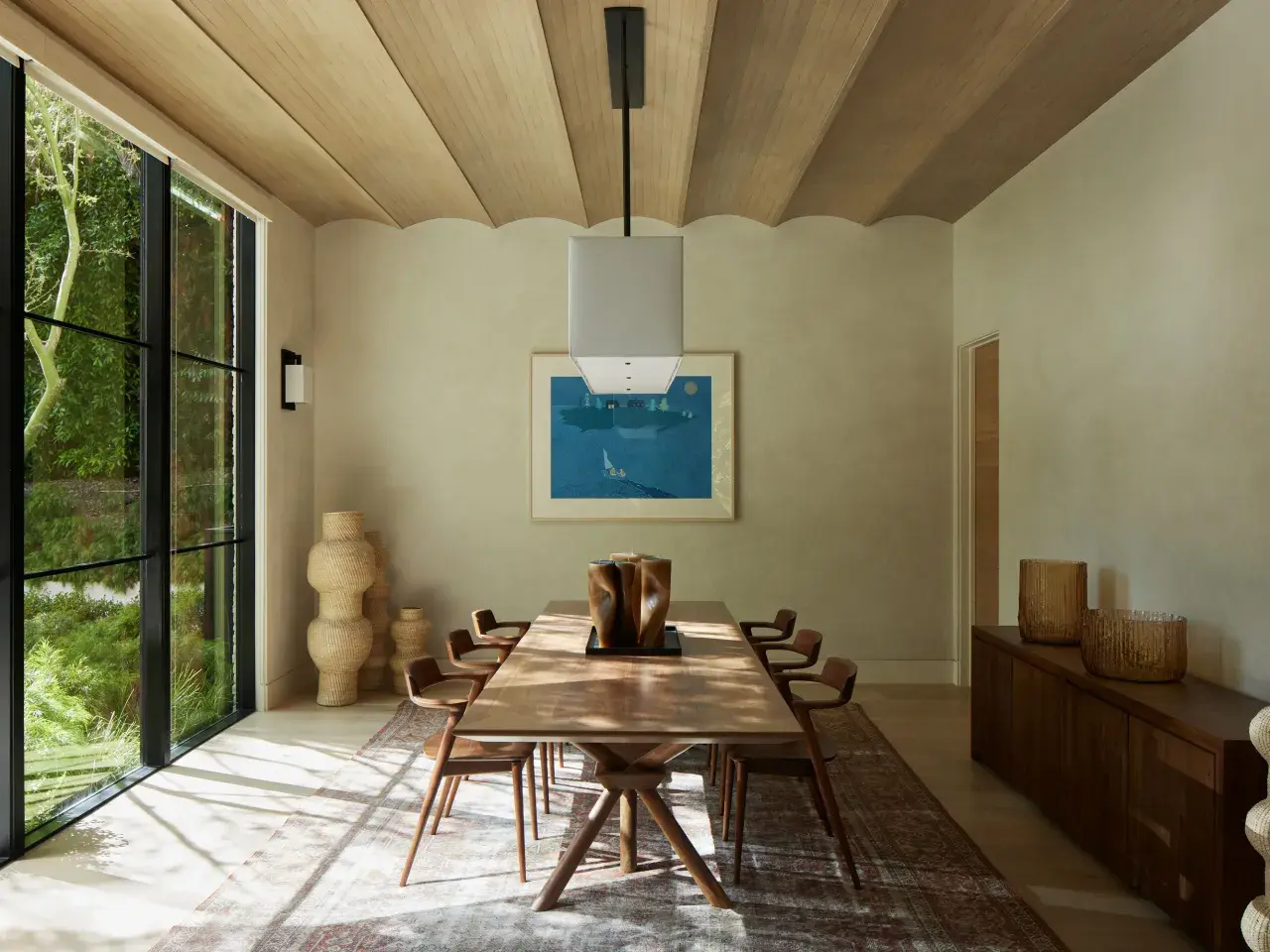 Comedor con mesa de madera, sillas de diseño y grandes ventanales. La decoración relajante se complementa con jarrones de mimbre y una pintura de paisaje marino.