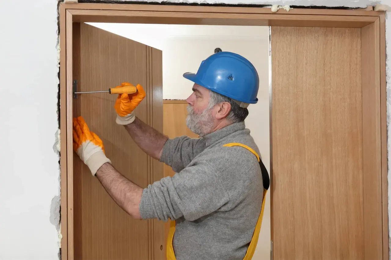 Hombre con casco azul y guantes naranjas ajustando una puerta interior de madera con un destornillador.