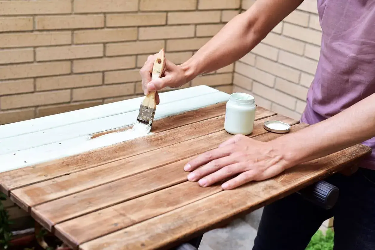 Persona pinta una mesa de madera barnizada con brocha y pintura blanca.