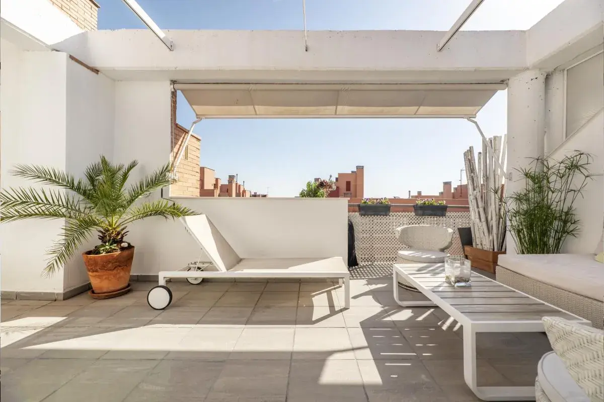 Ideas para decorar un ático: tumbona blanca, mesa de centro, sillas de mimbre y plantas verdes en una terraza soleada.