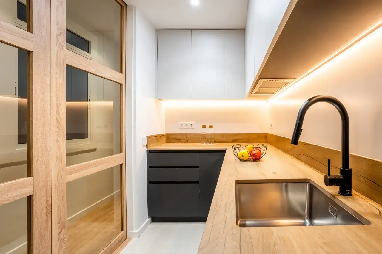 Cocina moderna con puertas de madera y cristal, encimera de madera, fregadero de acero inoxidable y grifo negro. Perfecta para reformas de cocinas pequeñas.