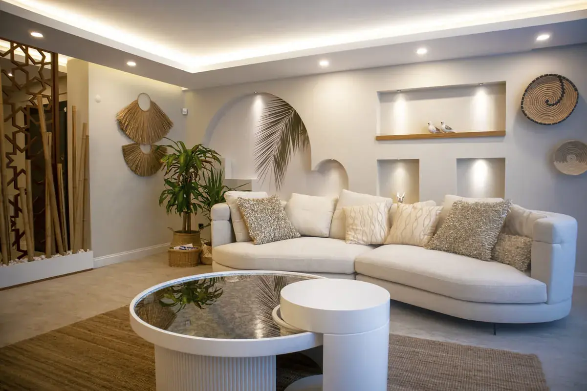 Sofá curvo blanco con cojines texturizados, mesa de centro redonda, y **últimas tendencias iluminación salones** con luces empotradas y perimetrales.