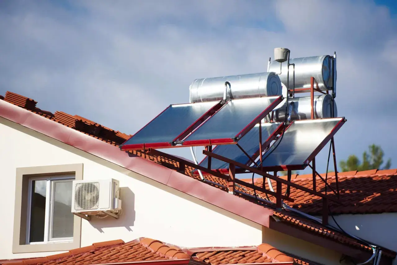 Paneles solares y tanques para agua caliente solar en un tejado de tejas rojas, bajo un cielo azul con nubes.