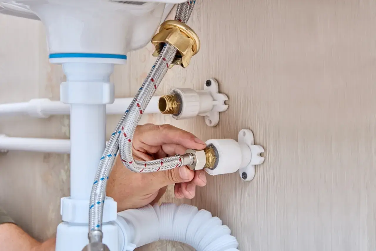 Mano ajustando la conexión de una manguera a la llave de paso de agua general, parte de la fontanería de un lavabo.