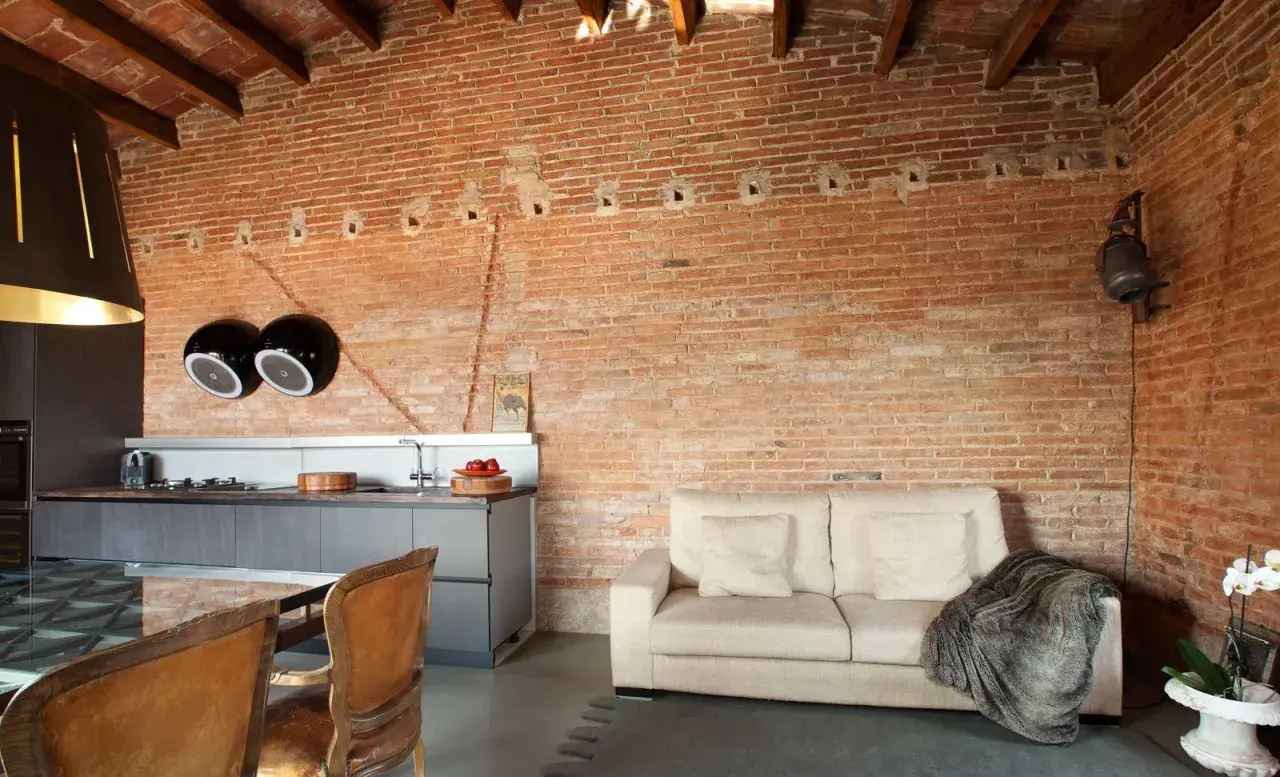 Un salón moderno con una pared rústica interior de ladrillo visto, sofá beige, cocina integrada y sillas de cuero.