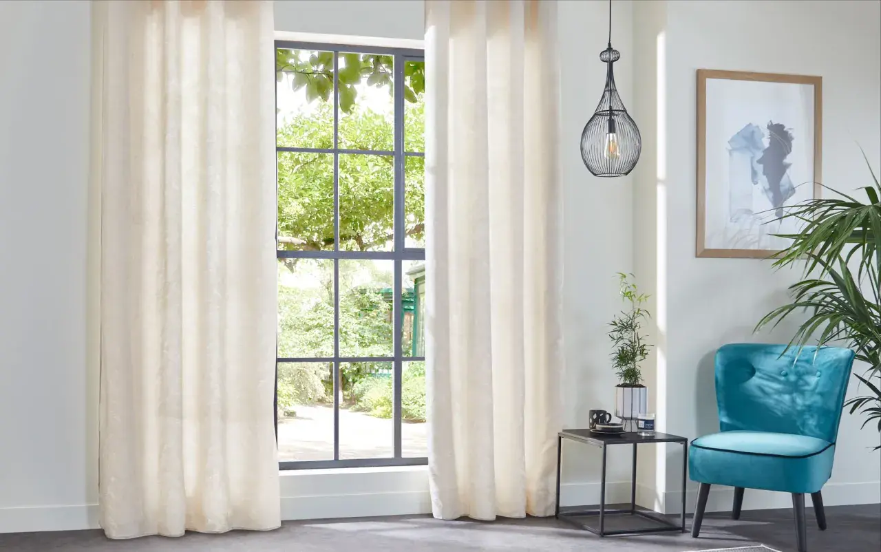 Elegantes cortinas de tela crema con sutil estampado, ideales para distintos tipos de cortinas para ventanas, complementan un sillón turquesa y un cuadro abstracto.