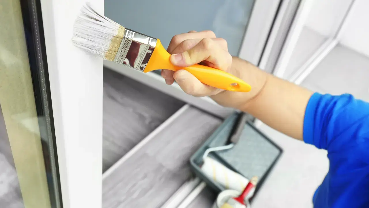 Mano pintando un marco de ventana blanco con una brocha. Se ve una bandeja de pintura y un rodillo en el fondo. Ideal para pintura para pintar plástico.