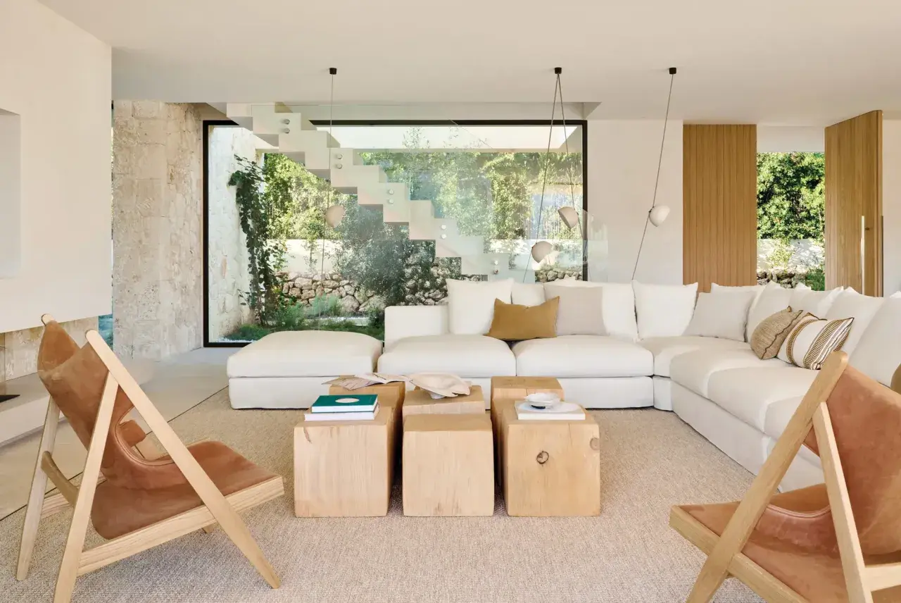 Amplio salón estilo mediterráneo con sofá blanco, mesas de madera y sillas de cuero, con vistas a un jardín exuberante a través de una escalera de cristal.