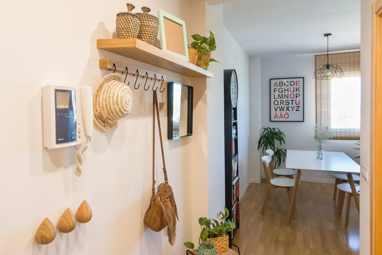 Pasillos con madera en la pared, un perchero con sombrero y bolso, y un intercomunicador. Al fondo, un comedor con mesa y sillas.