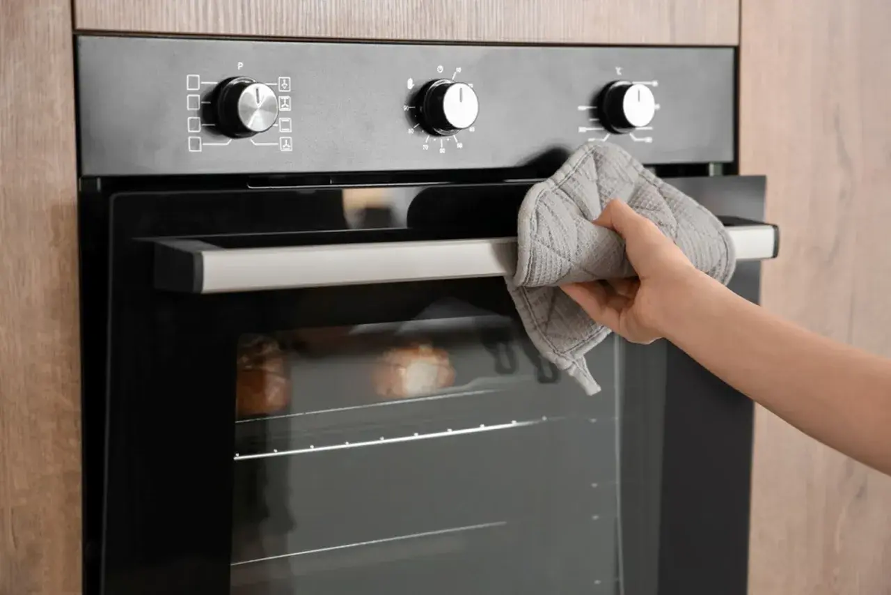 Mano con manopla sujetando la puerta del horno, donde se ve una sección de cable para horno y pan horneándose.