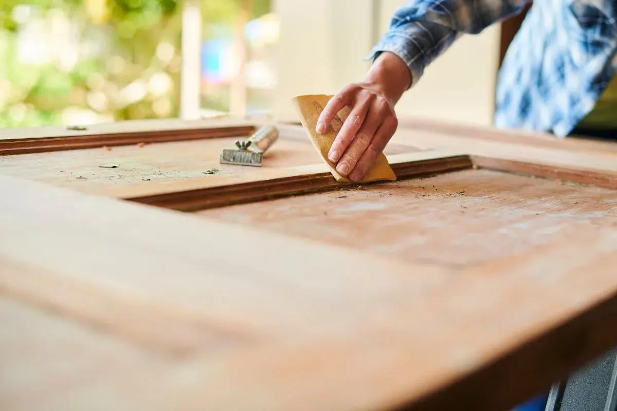 Mano lijando una puerta de madera exterior para restaurarla. Se ve el raspador al lado.