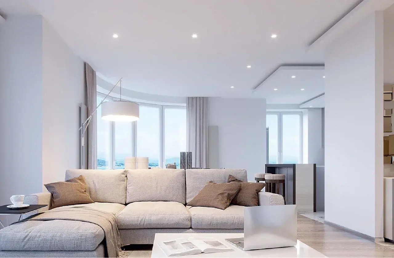 Salón moderno con sofá beige, mesa de centro y un techo pladur con focos empotrados. Ventanales curvos con vistas al mar.