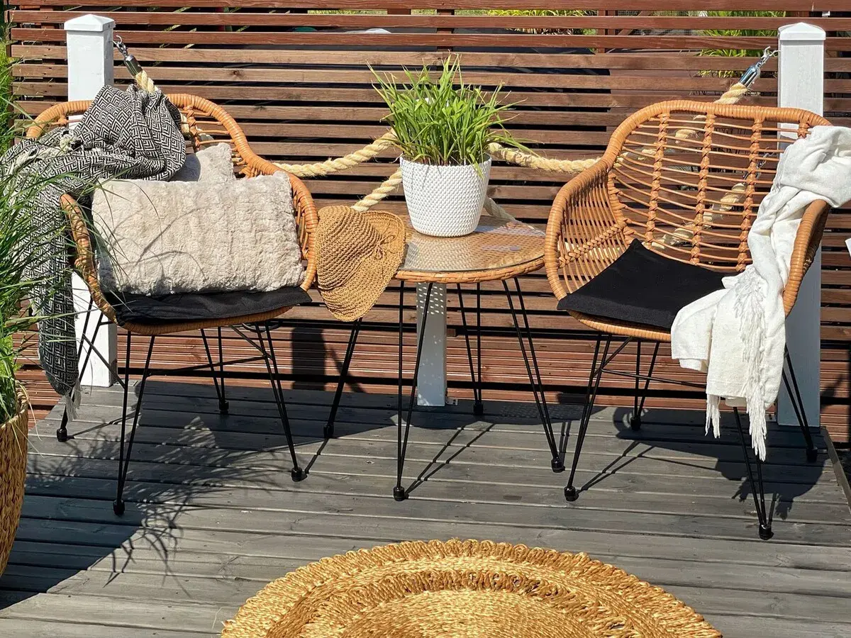 Dos sillas de mimbre con cojines y mantas, una mesa auxiliar con planta y un sombrero de paja. Decoración de mini balcones acogedora.
