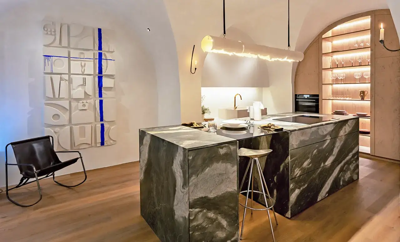 Elegante cocina con isla de m&aacute;rmol oscuro, arte abstracto en la pared y una silla de cuero.