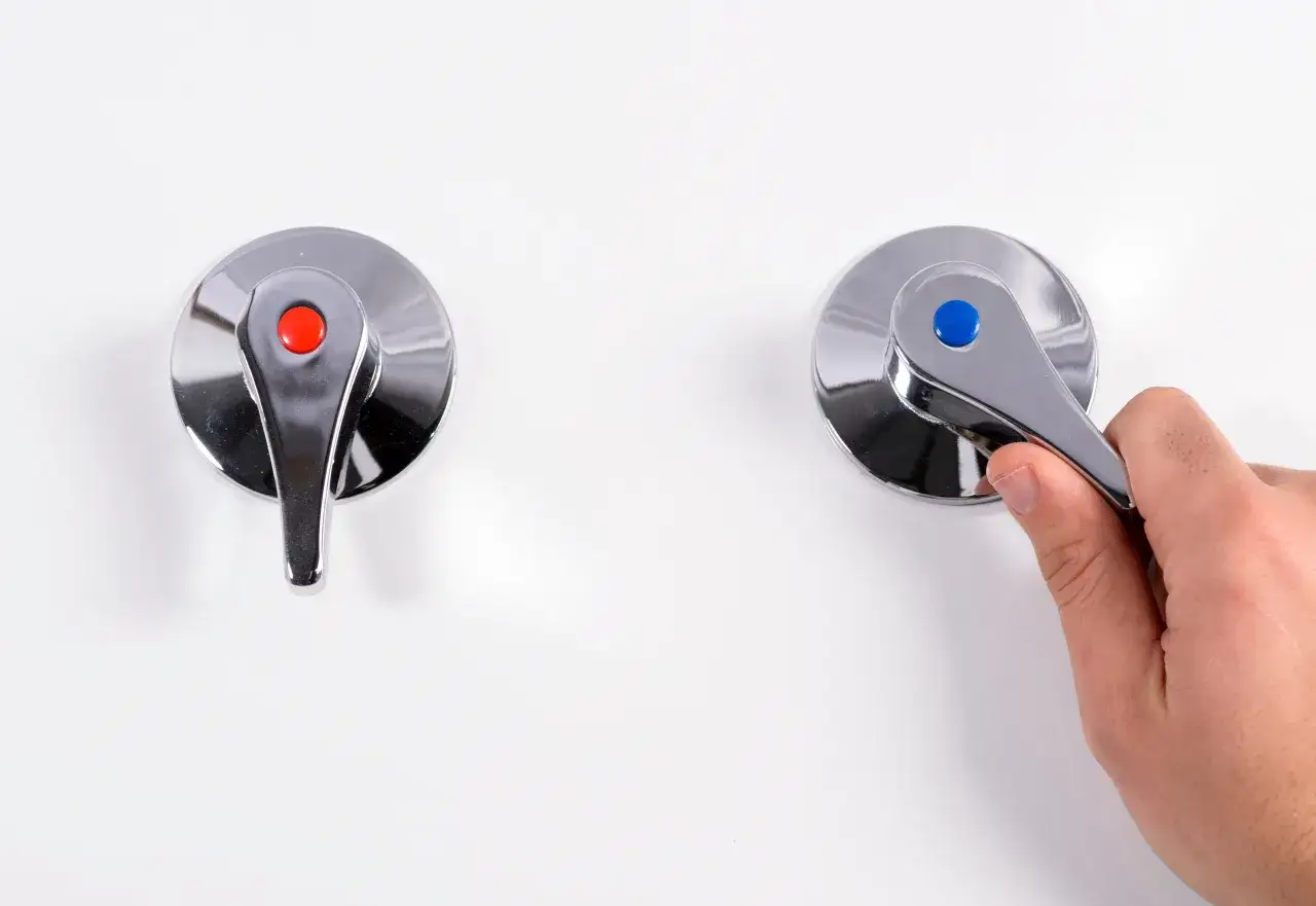 Dos grifos cromados, uno con bot&oacute;n rojo y otro con bot&oacute;n azul, sobre fondo blanco. Una mano gira el grifo azul.