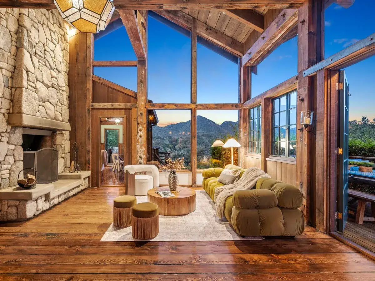 Acogedora sala con chimenea de piedra y vigas de madera, perfecta para una decoracion casa rustica. Sofá verde y vistas a la montaña.