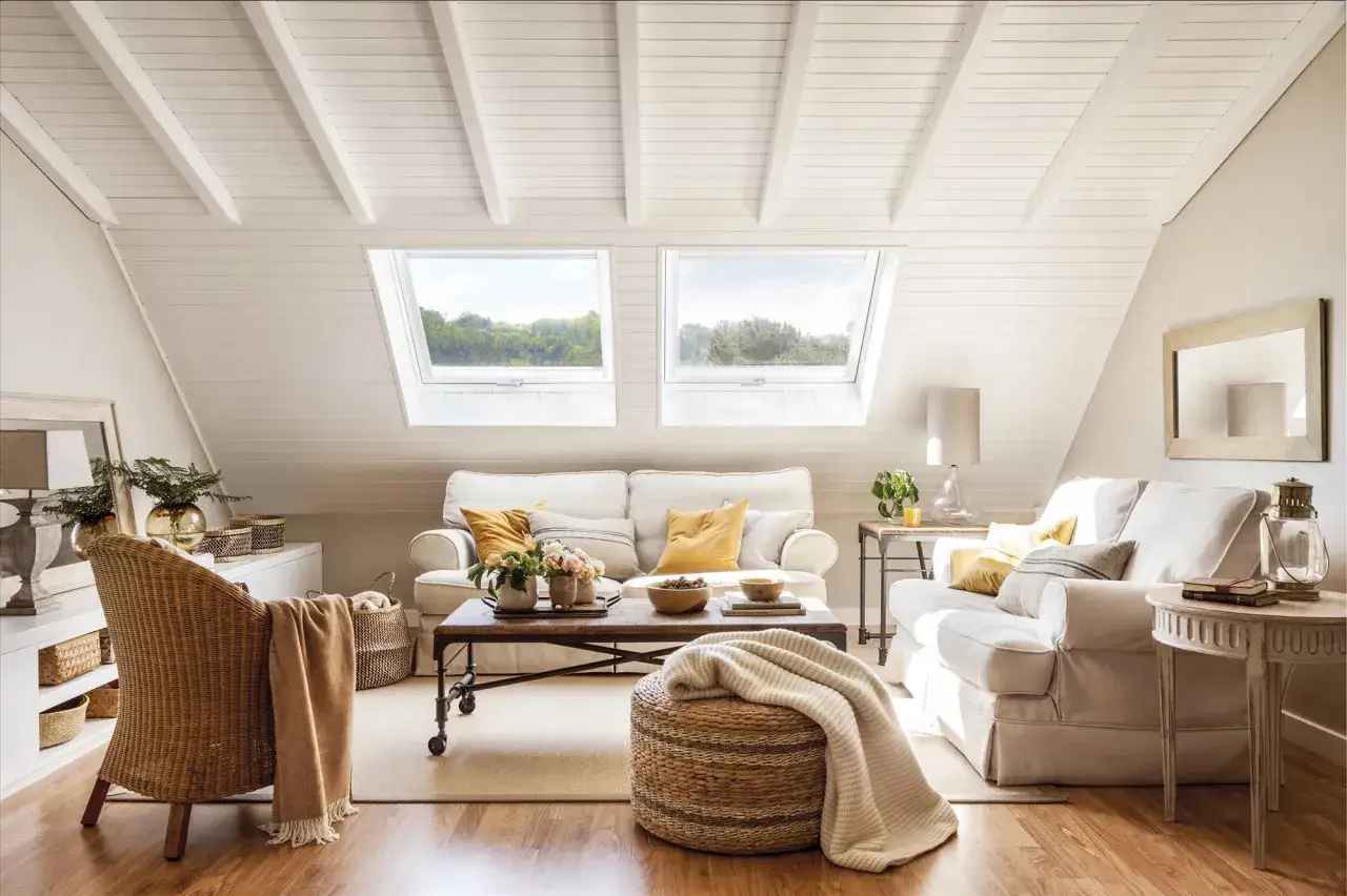 Ideas para decorar un &aacute;tico: sala de estar luminosa con sof&aacute;s blancos, mesa de centro de madera, sill&oacute;n de mimbre y ventanas en el techo.