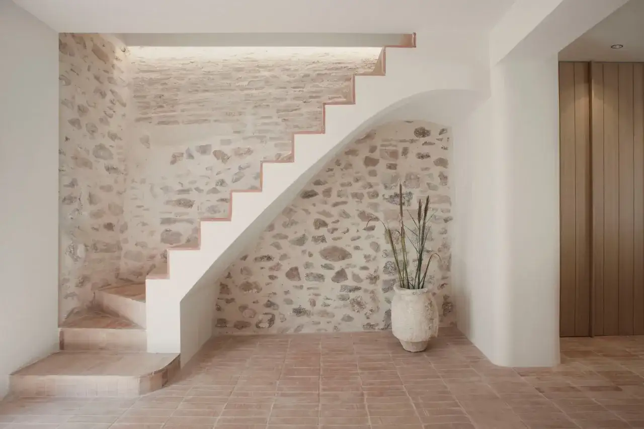Elegante subida de escalera interior con paredes de piedra rústica y escalones blancos. Un jarrón con plantas decora el espacio, creando un ambiente acogedor.