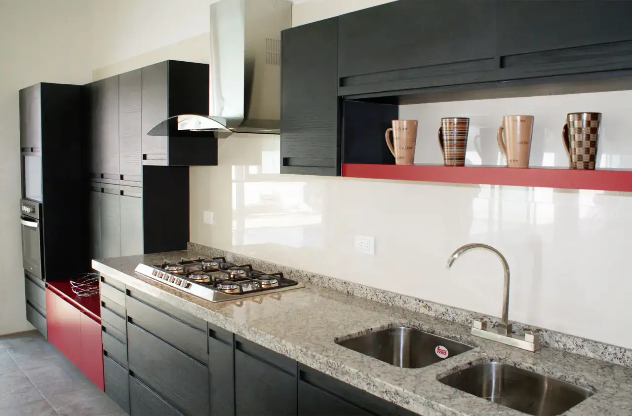 Cocina moderna con gabinetes oscuros, encimera de granito, estufa, campana extractora y tazas en una repisa roja, siguiendo las normas de la cocina.