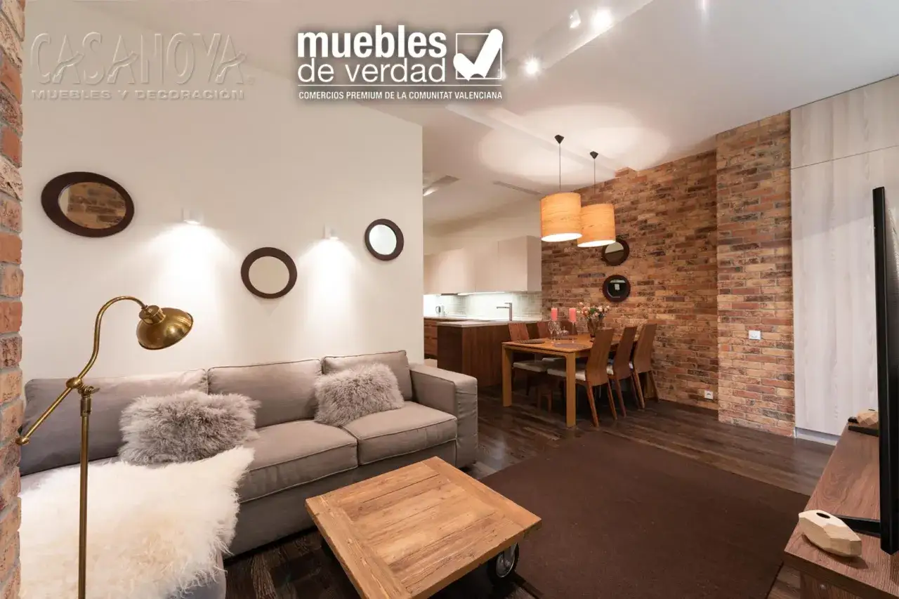 Sof&aacute; gris, mesa de centro de madera y l&aacute;mpara dorada, ideal para aprovechar espacios peque&ntilde;os. Comedor con mesa de madera y sillas, junto a pared de ladrillo.