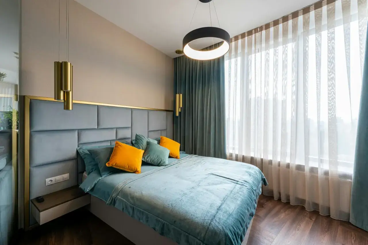 Cabecero tapizado gris con detalles dorados, ideal para tus ideas para hacer un cabecero. Cama con ropa de cama azul y cojines naranjas.