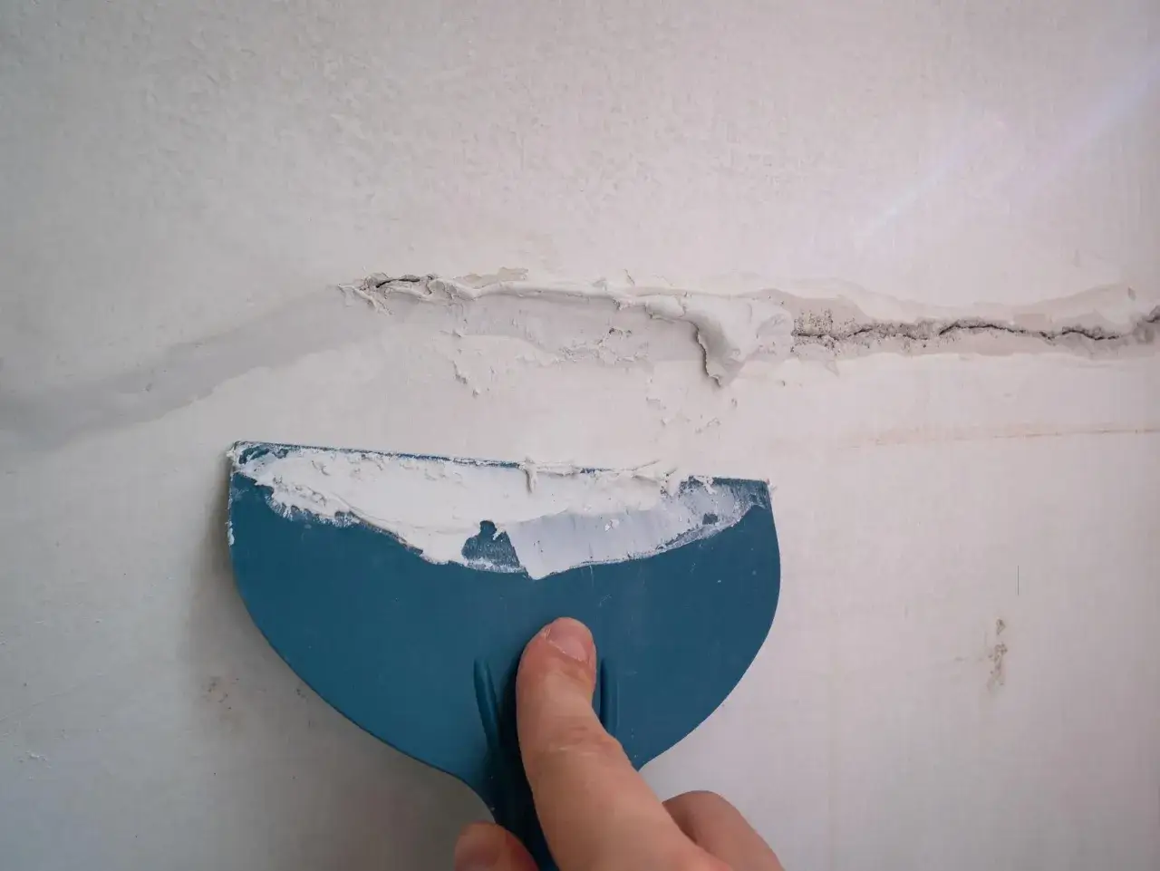 Una mano usa una esp&aacute;tula azul para aplicar masilla sobre una grieta en una pared, prepar&aacute;ndola para lijar paredes.