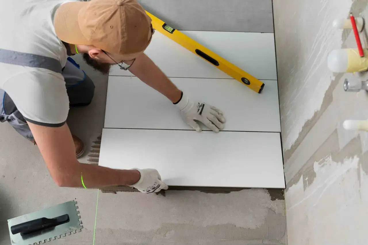 Un hombre con guantes y gorra coloca azulejos blancos en el suelo, usando un nivel y un láser para asegurar la precisión. Aprende como poner azulejos.