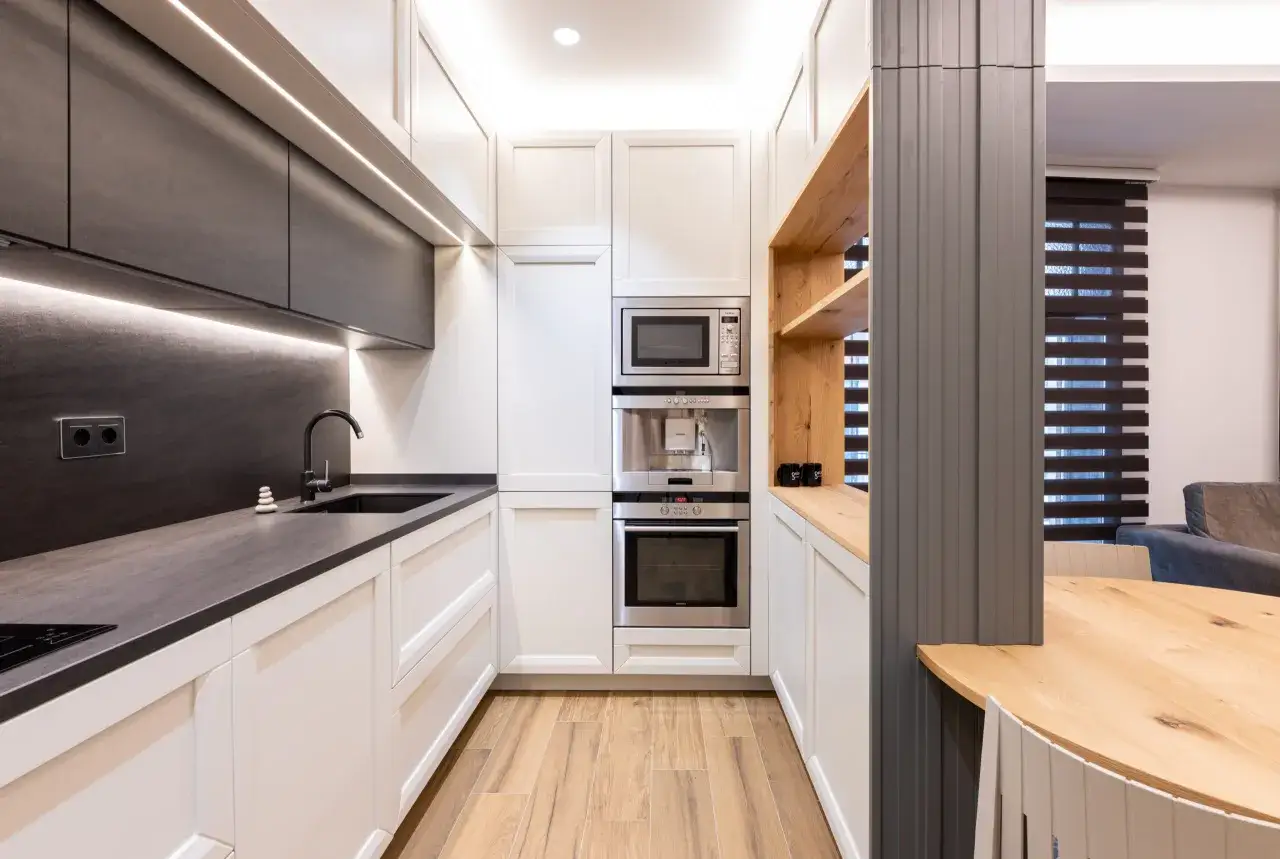 Moderna cocina pequeña con gabinetes blancos y oscuros, encimeras de piedra, electrodomésticos integrados y suelo de madera.