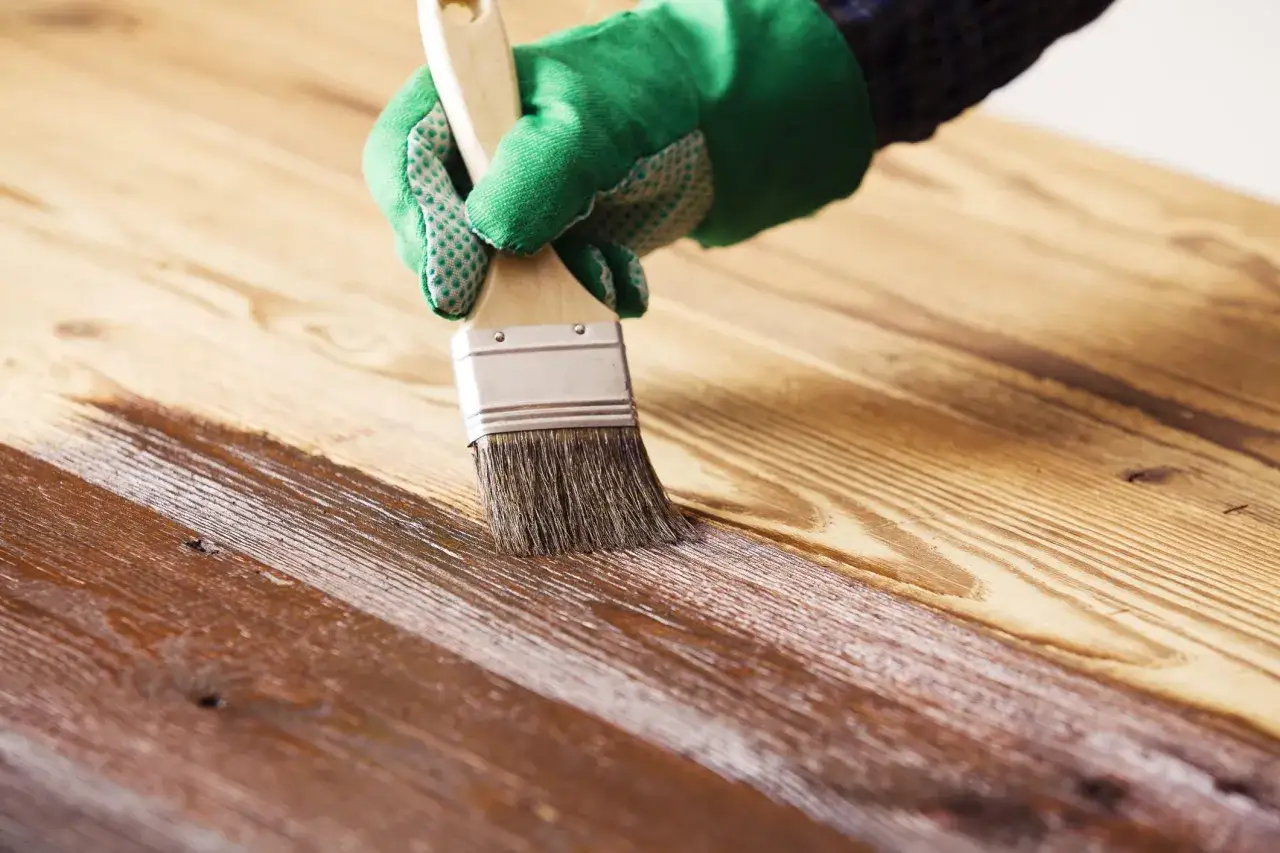 Mano con guante verde aplicando barniz sobre madera, creando una capa protectora. Barnizar sobre barniz sin lijar para un acabado r&aacute;pido.
