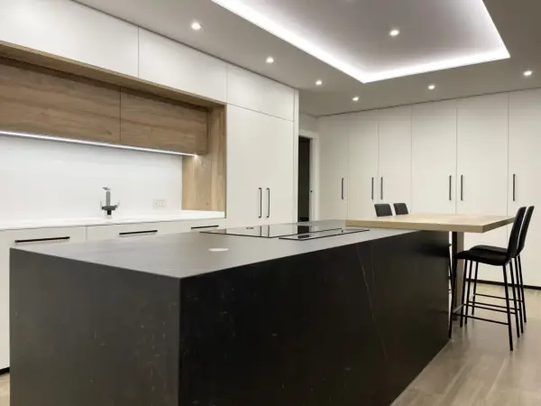 Cocina moderna con isla negra, encimeras blancas y armarios de madera. La iluminación LED bajo los armarios y en el techo resalta el diseño, mostrando cómo iluminar una cocina con estilo.